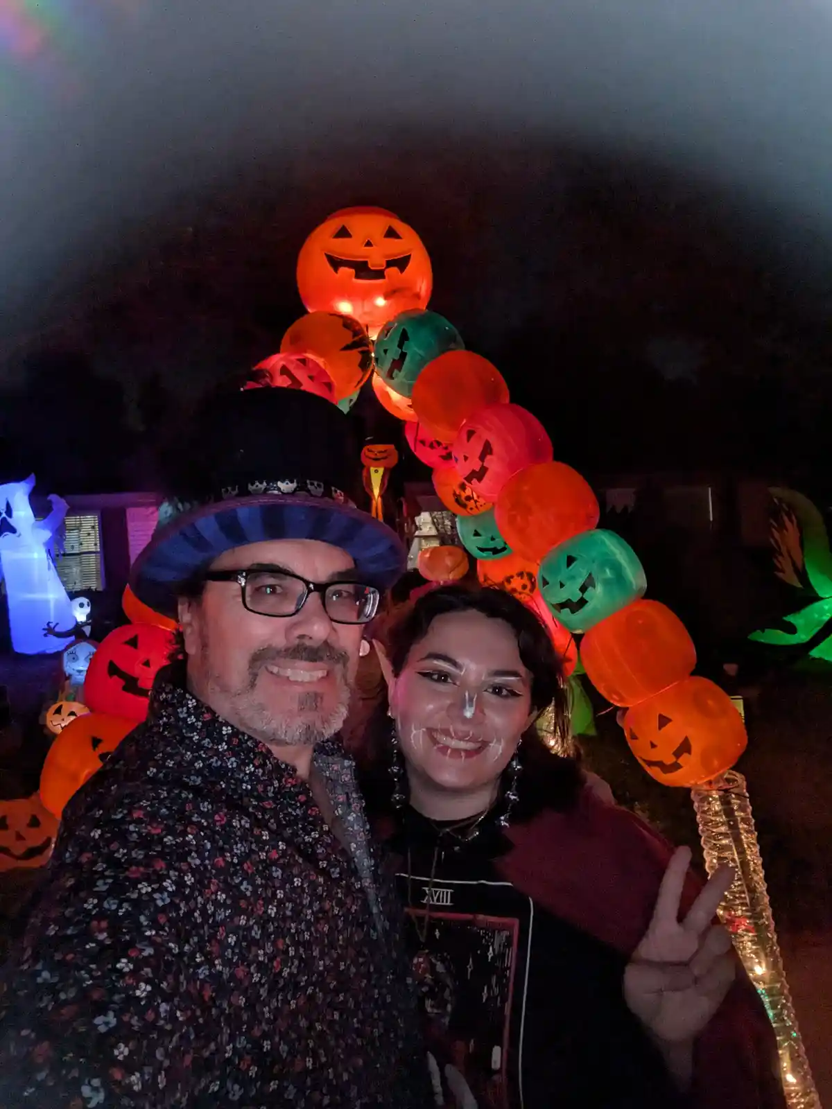 A man and woman pose for a selfie at night in front of a large arch made of glowing jack-o'-lantern balloons. The man wears…