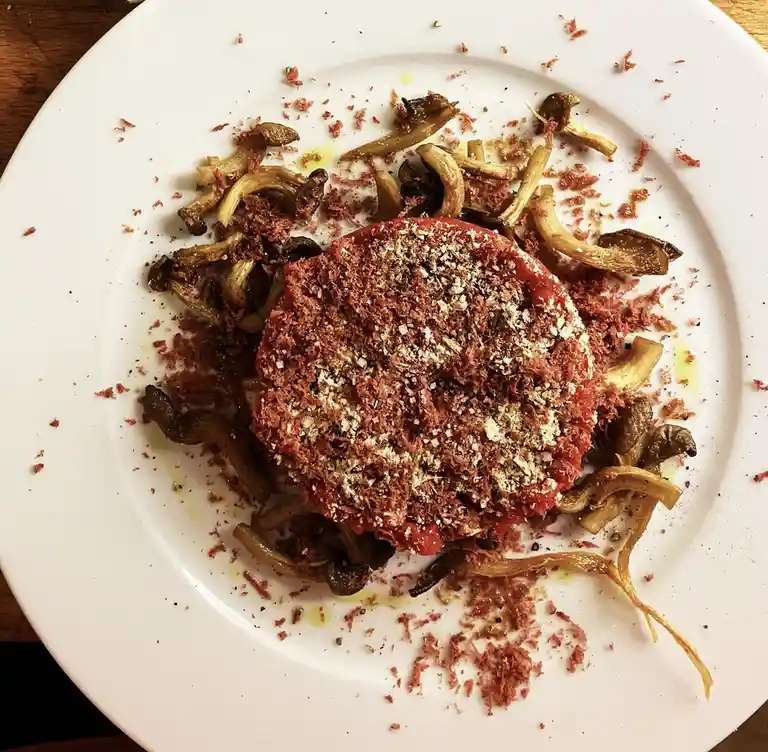 A white plate holds a dish that appears to be a raw meat tartare, topped with coarse salt and pepper, surrounded by roasted…