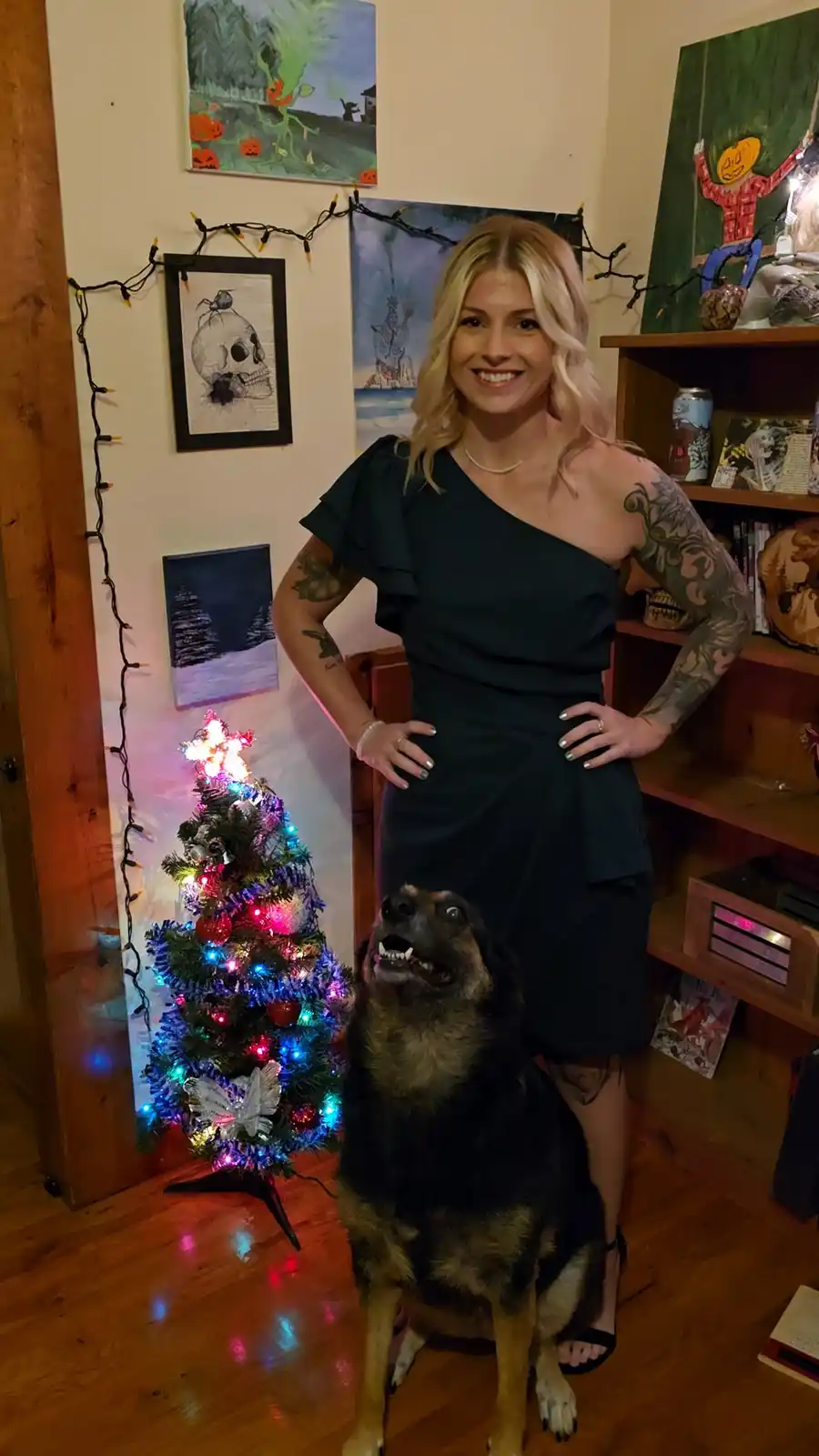A woman with blonde hair and tattoos stands confidently with hands on hips next to a German Shepherd dog that is looking up…