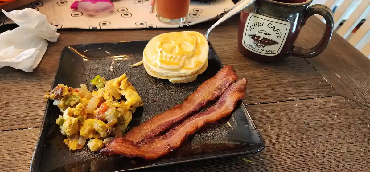 A breakfast plate with two strips of bacon, scrambled eggs with vegetables, and a stack of pancakes shaped like a skull, all…