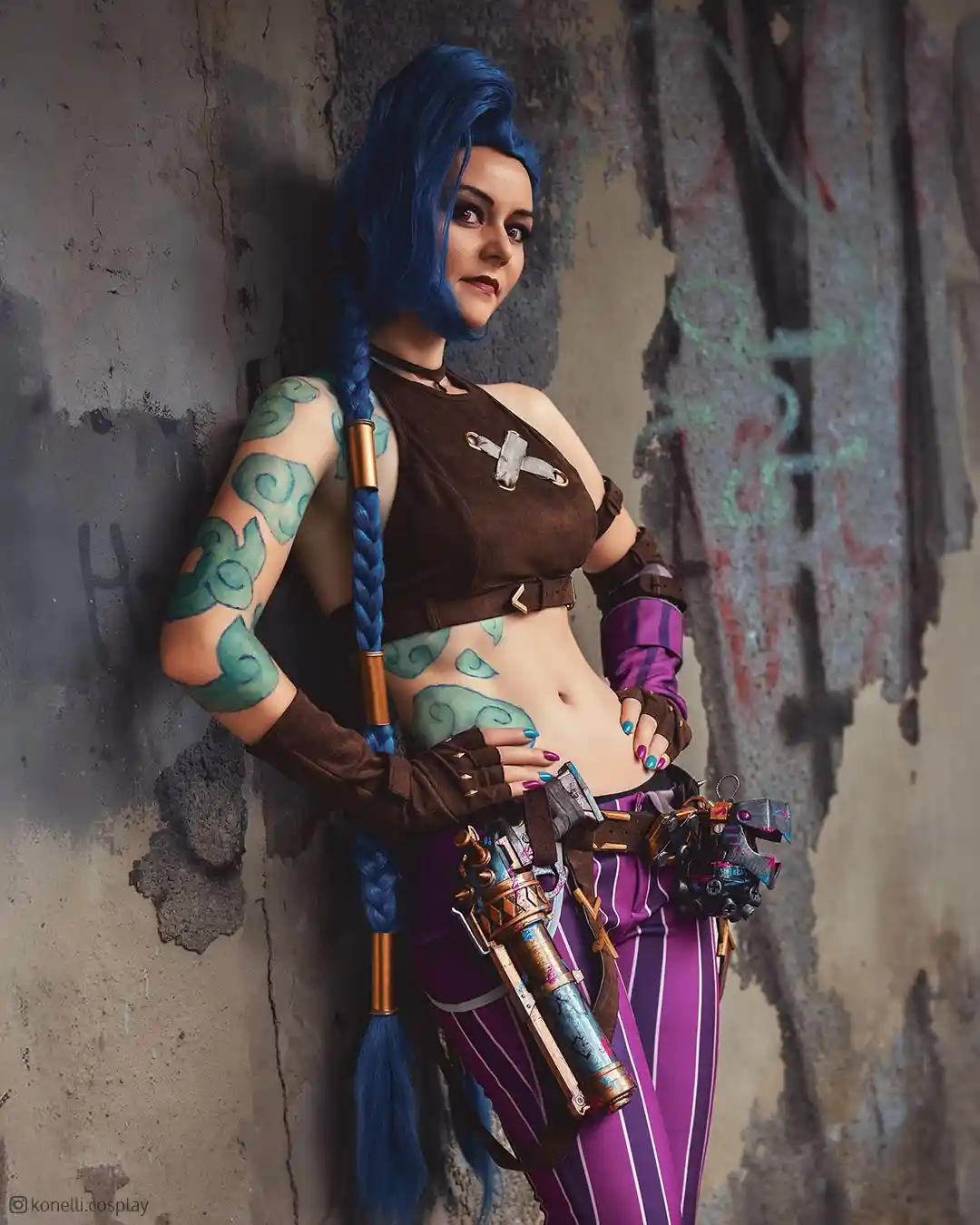 A person in a detailed cosplay of the video game character Jinx, featuring bright blue braided hair, green cloud tattoos, a…