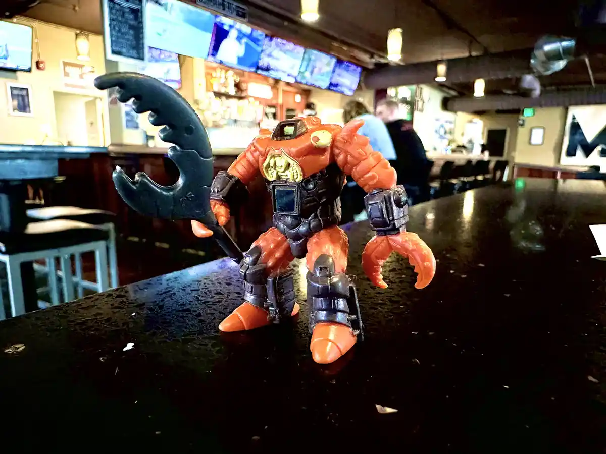 An orange and black toy robot with claw-like hands and a large black scythe stands on a dark bar counter in a dimly lit bar…