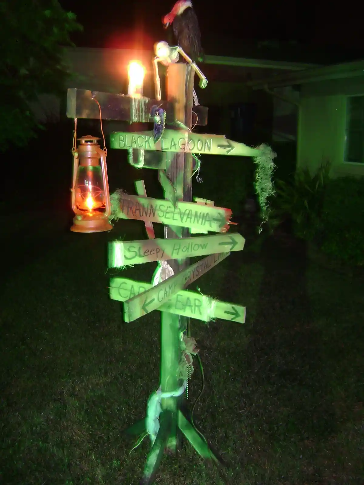 A spooky, homemade Halloween signpost at night, decorated with a glowing lantern, a skeleton, and a vulture, with…