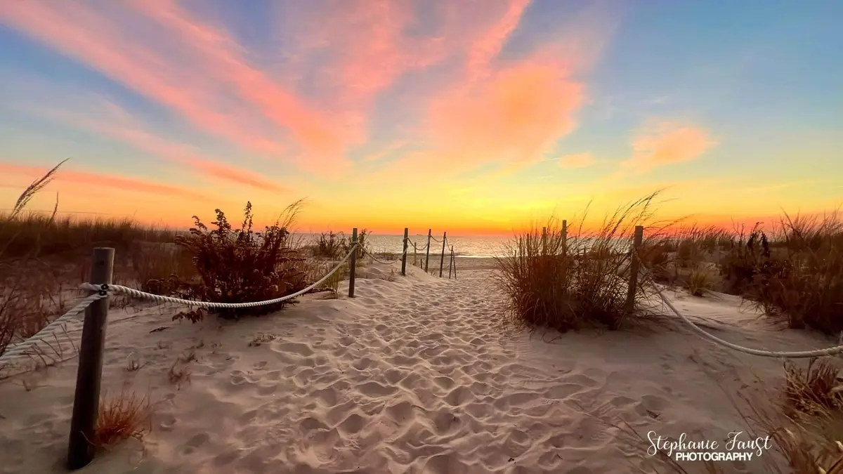 A serene beach sunset photo shows a sandy path bordered by rope fences leading to the ocean, with vibrant pink, orange, and…