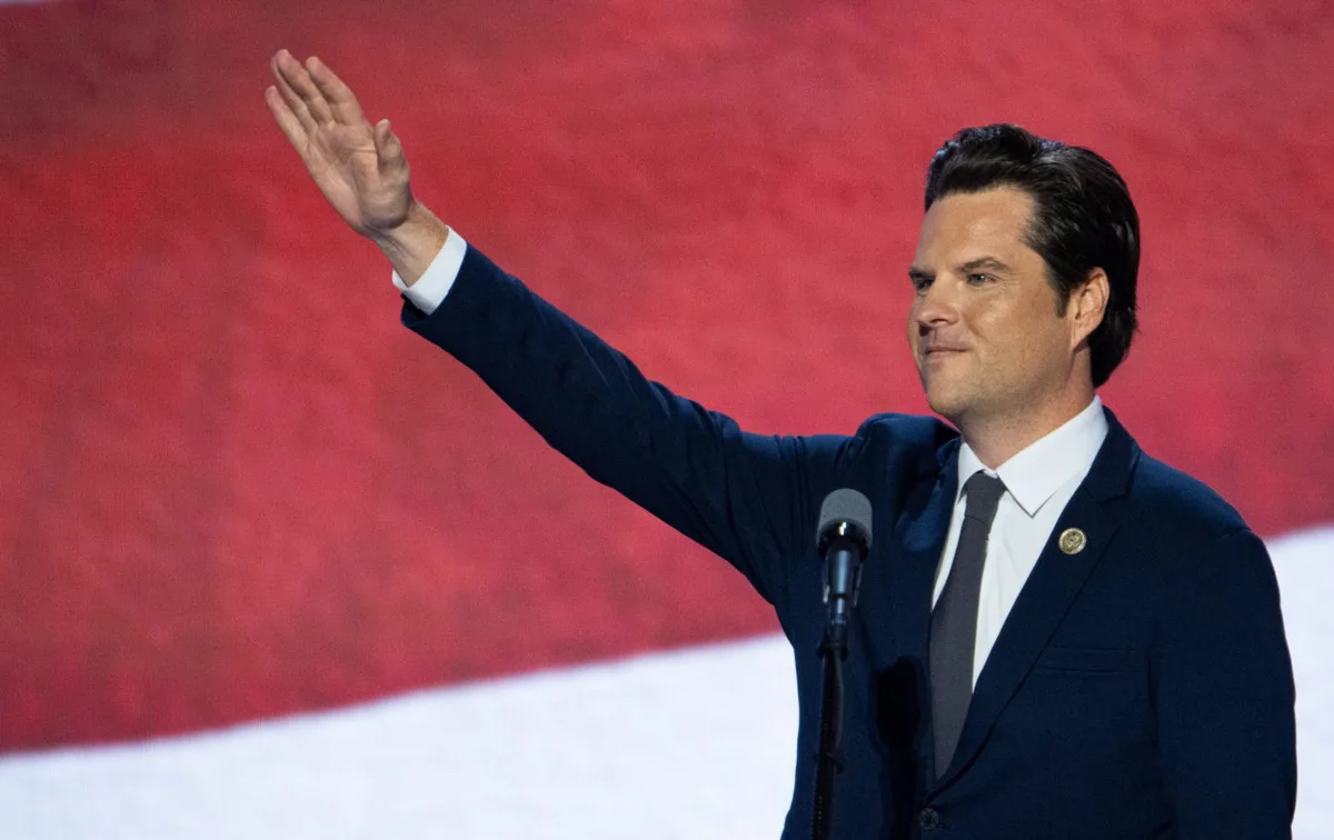 a man in a dark suit and tie stands at a podium with a microphone, waving his right hand while smiling slightly. Behind him…