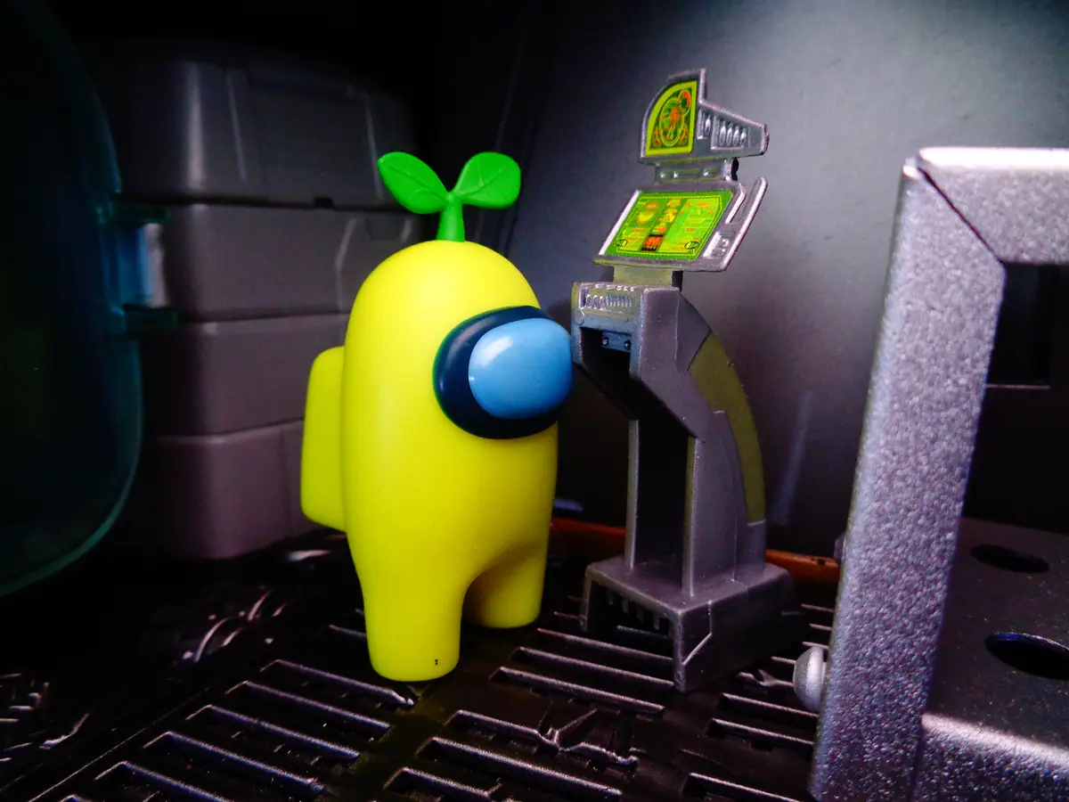 A yellow Among Us crewmate figurine with a small green sprout growing from its head stands next to a slot machine in a dimly…