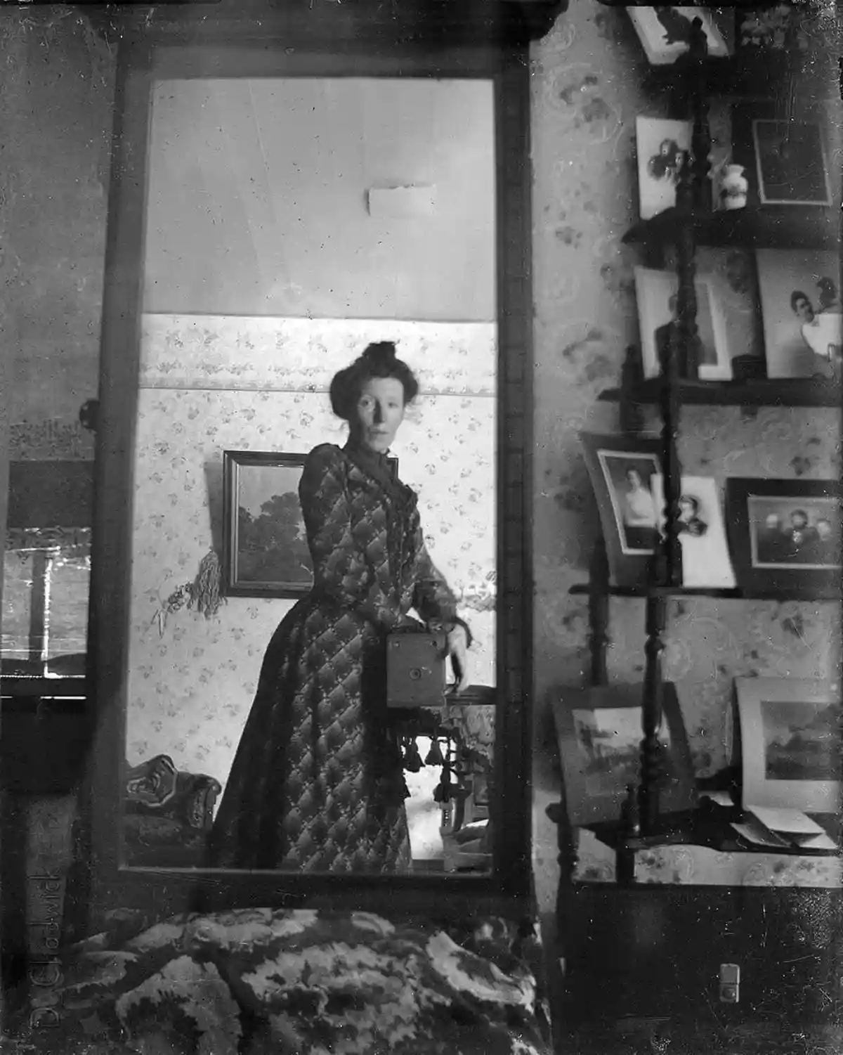 A vintage black-and-white photograph shows a woman in a patterned dress standing in front of a mirror, holding an…
