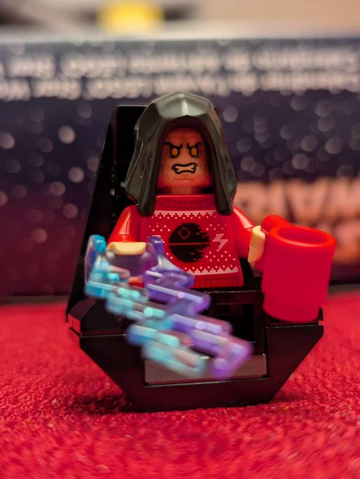A LEGO minifigure resembling Darth Vader sits in a black chair, wearing a red Christmas sweater with a Death Star and…