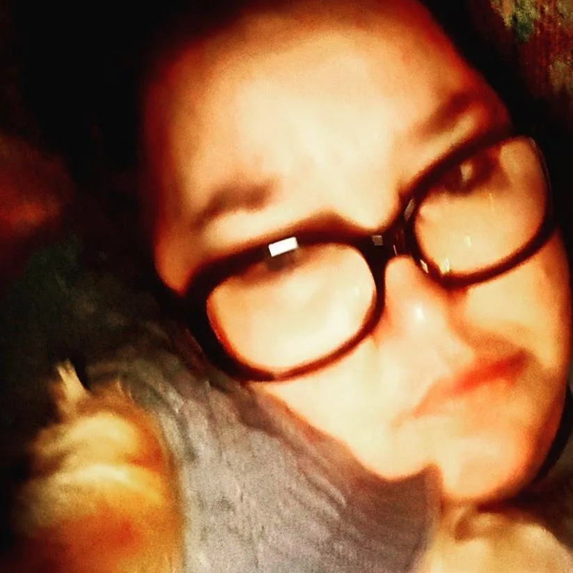 A blurry, low-resolution selfie of a person wearing thick-rimmed glasses, looking directly at the camera with a neutral or…