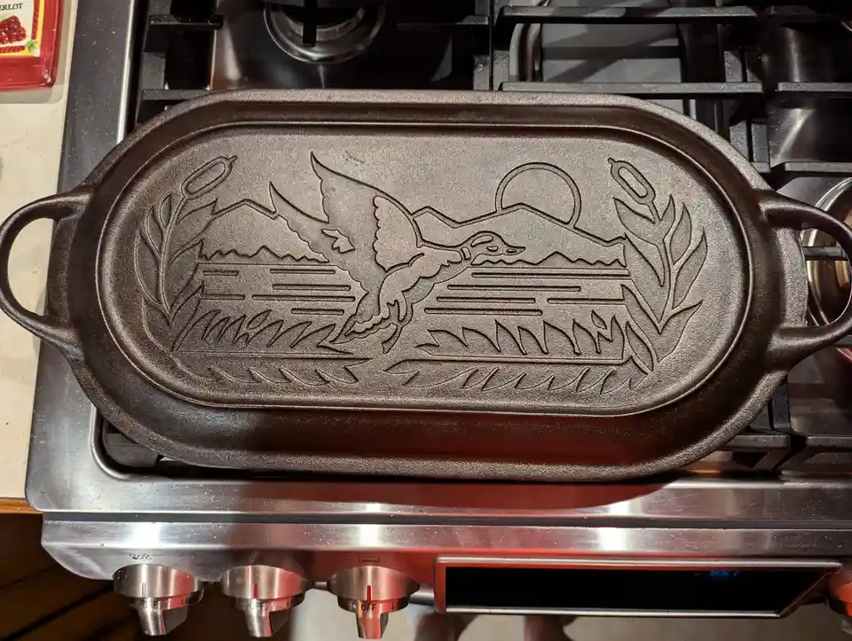 A dark cast iron skillet with a detailed embossed design of a duck flying over water with reeds and a sun is placed on a…