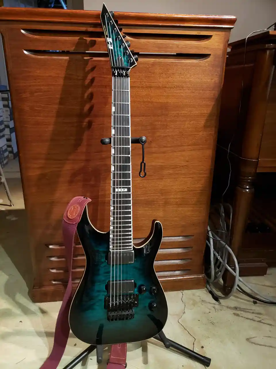 A teal and black electric guitar with a red strap is propped up on a stand in front of a wooden cabinet. The guitar has a…
