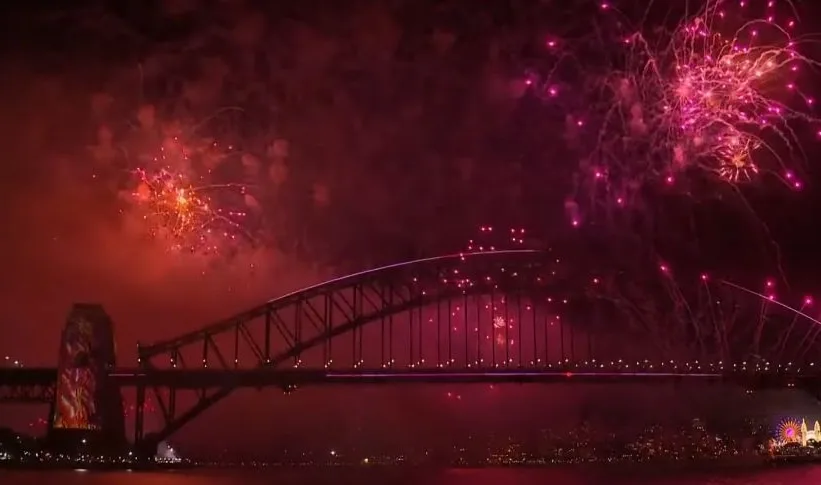 A nighttime view of the Sydney Harbour Bridge illuminated with lights, with vibrant red and pink fireworks exploding in the…
