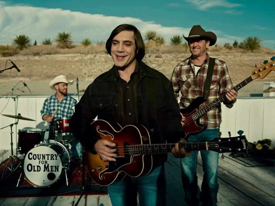 Three men in cowboy attire play instruments outdoors on a wooden stage with a desert backdrop; the bass drum in front reads…