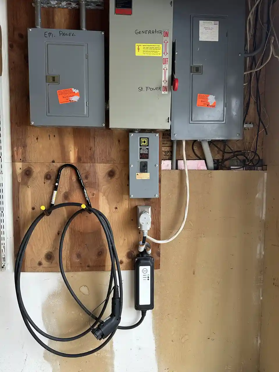 Three gray electrical panels mounted on a wooden wall, labeled 'EM. PANEL', 'GENERATOR', and 'St. POWER', with a coiled…