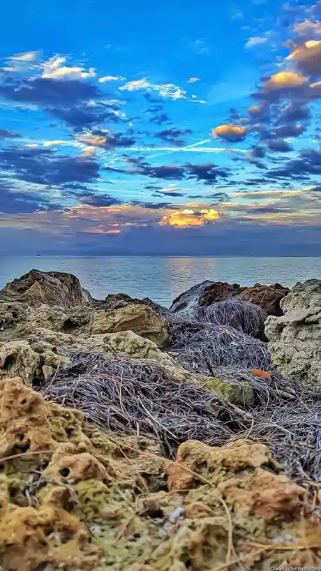 A scenic photo of a rocky shoreline at sunset or sunrise, with vibrant blue and orange clouds above calm water. Piles of…