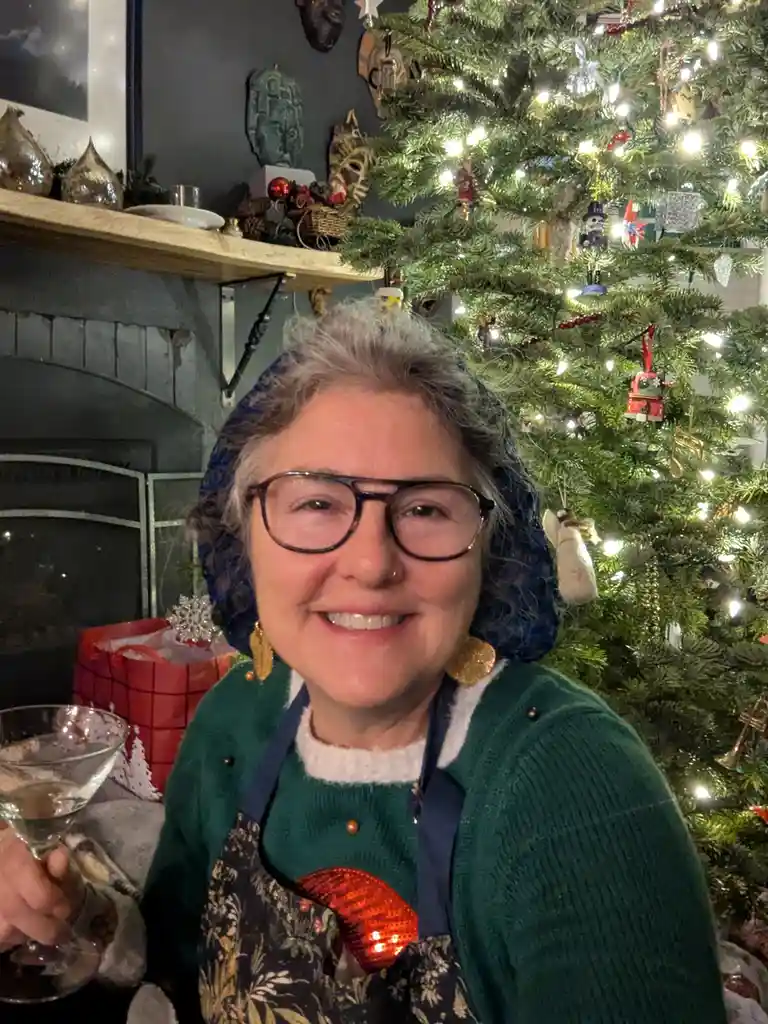 A smiling woman with glasses and a blue headscarf holds a martini glass in front of a decorated Christmas tree. She wears a…