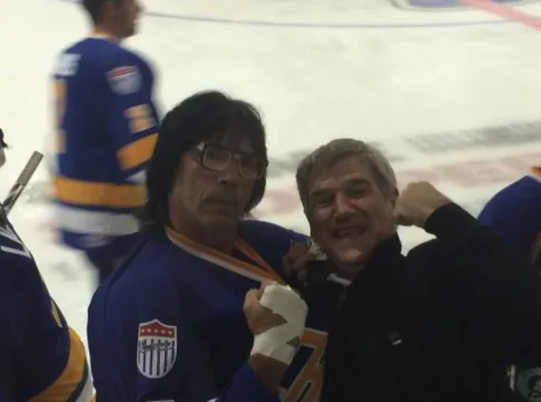 Two men are posing for a photo on an ice hockey rink. The man on the left, wearing a blue hockey jersey with an American…