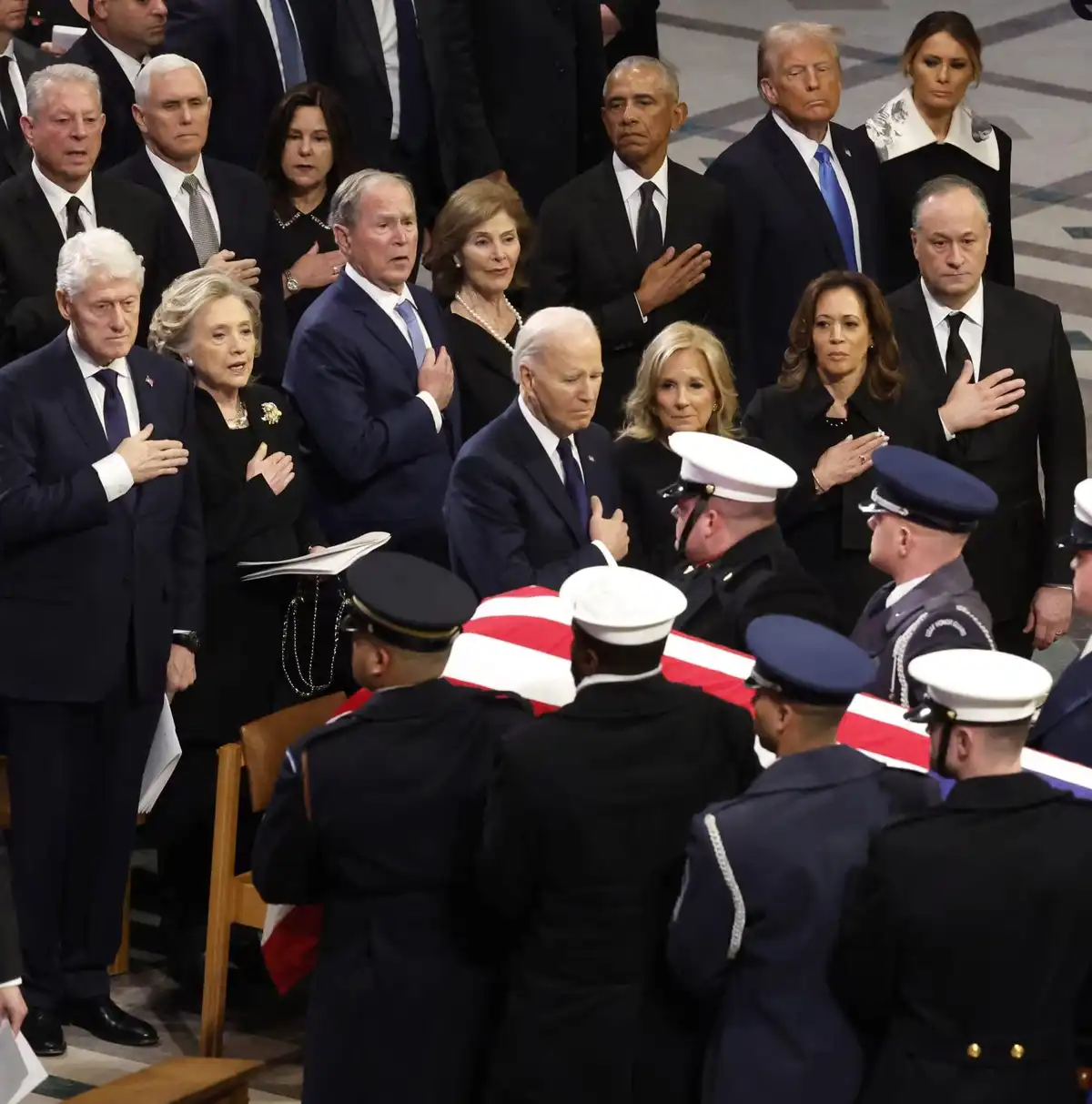 A solemn photo of former U.S. presidents and their families at a state funeral, standing in formal black attire with hands…