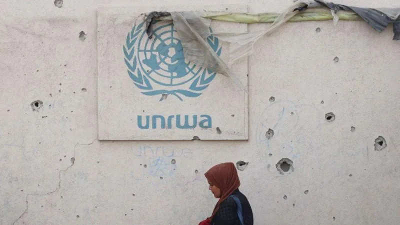 A weathered UNRWA logo is affixed to a bullet-riddled concrete wall, partially covered by torn plastic. A woman wearing a…