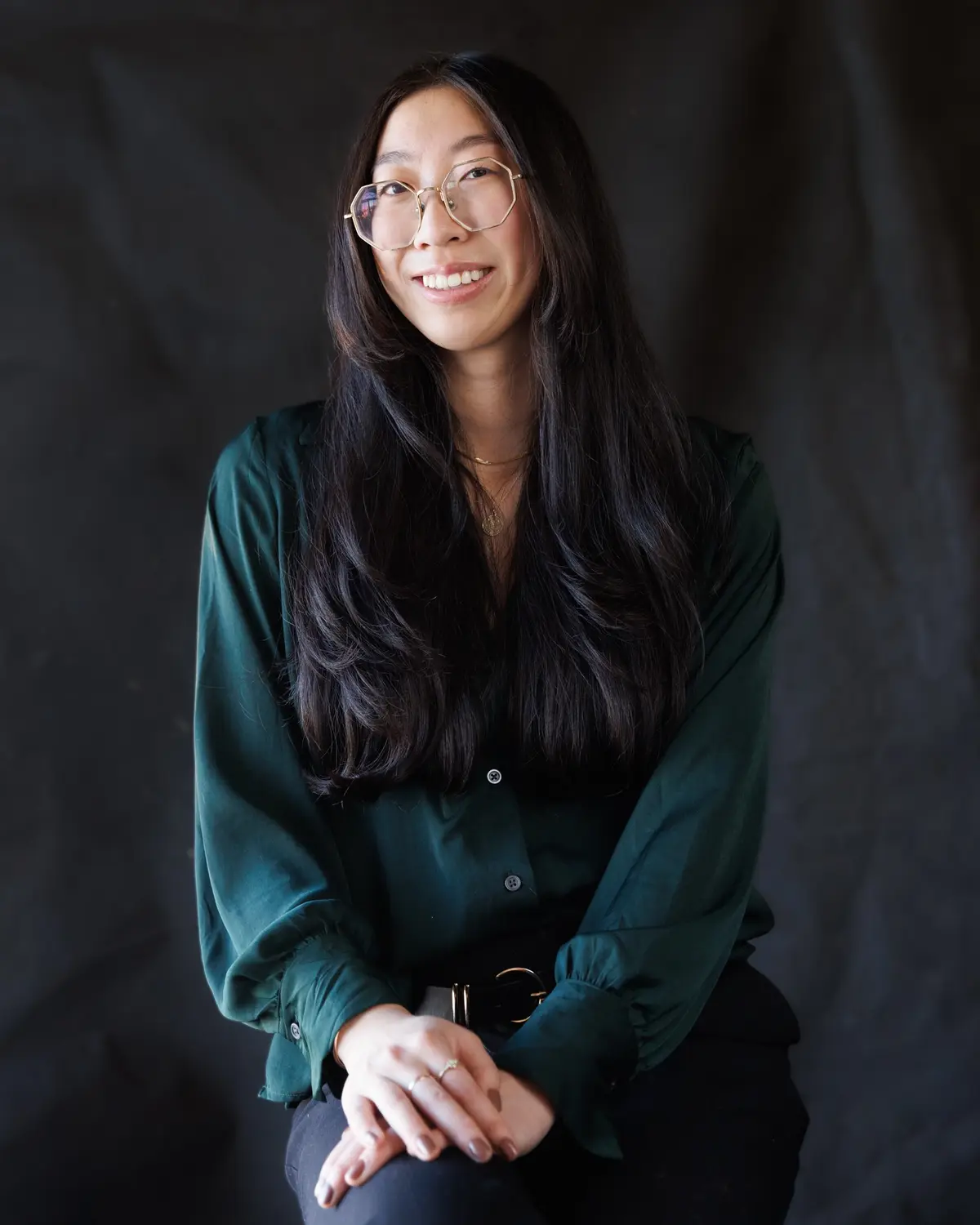 A woman with long dark hair and gold-rimmed glasses smiles warmly while seated against a dark backdrop, wearing a green…
