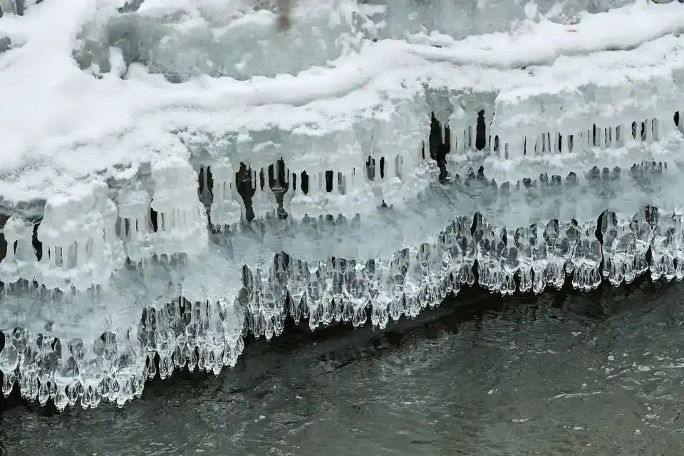 A close-up photograph shows layered ice formations with icicles hanging over dark water, topped with snow. The overlaid text…