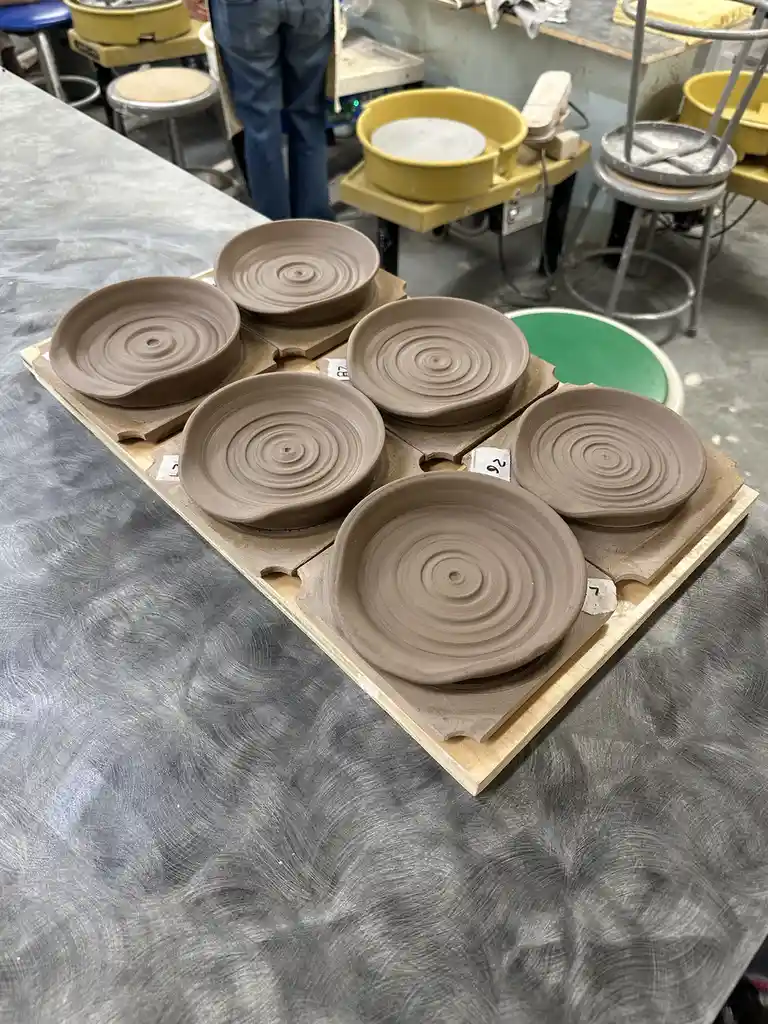 Six identical, unglazed clay bowls with concentric spiral patterns sit on a wooden board in a pottery studio, each labeled…