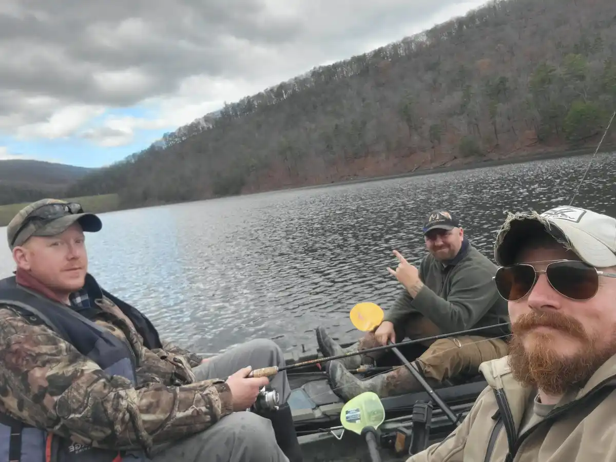 Three men are in a boat on a lake with forested hills in the background. The man in the foreground wearing sunglasses and a…