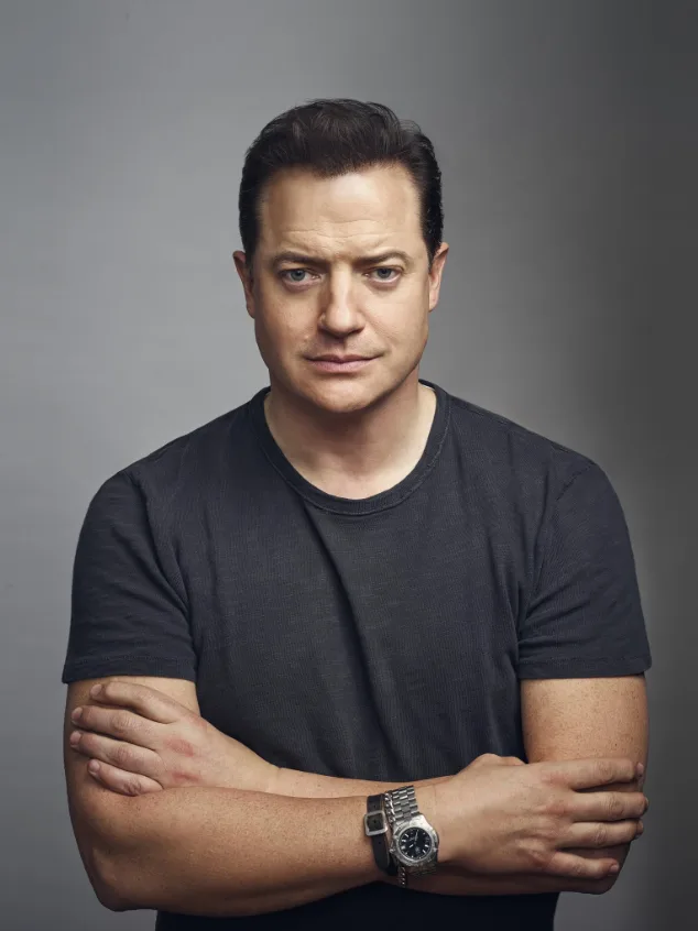 a man with dark hair and a serious expression stands with his arms crossed, wearing a dark t-shirt and a watch on his left…