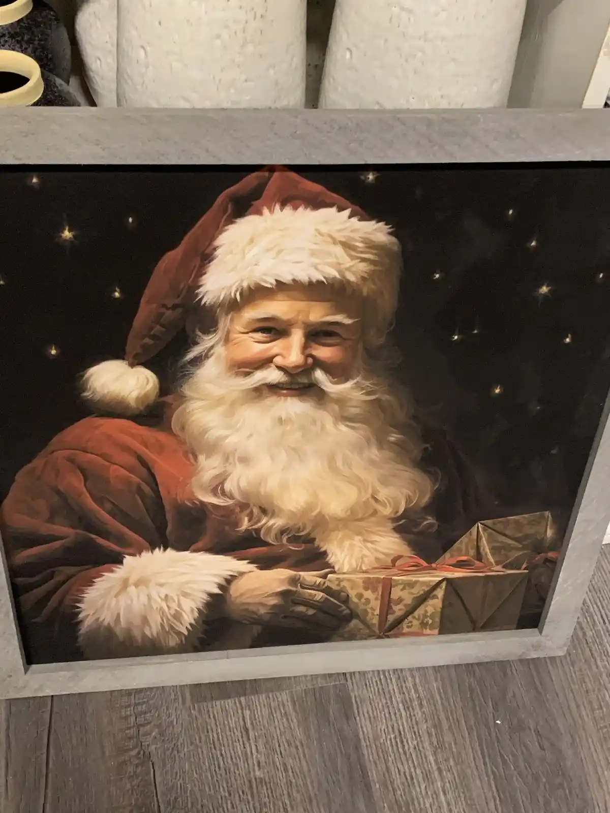 A framed painting of a smiling Santa Claus in a red suit with white fur trim, holding a wrapped gift against a starry night…