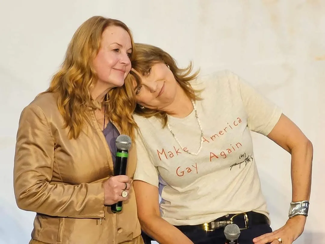 Two women are posing together on a stage; the woman on the right wears a white t-shirt with the phrase 'Make America Gay…