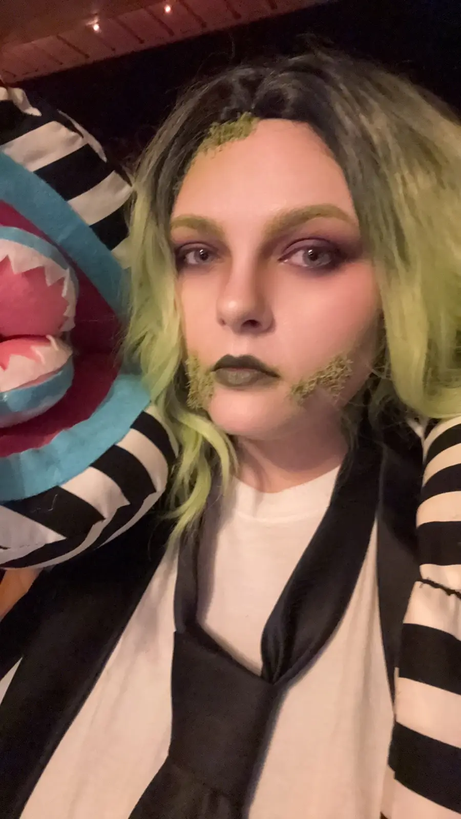 A person in a cosplay of the character Wednesday Addams, with green hair, dark makeup, and moss-like green substance on…