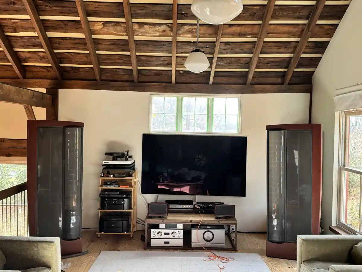 A room with a large TV flanked by two tall floor-standing speakers, surrounded by various audio equipment on shelves and a…