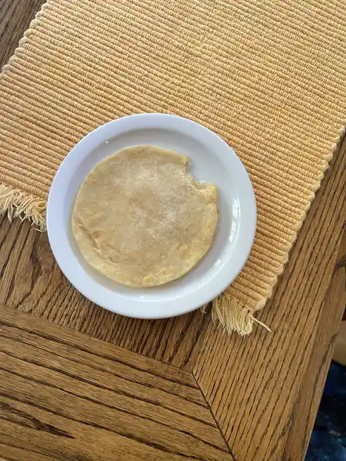 A single, partially eaten tortilla rests on a white plate, which sits atop a woven yellow placemat on a wooden table. The…