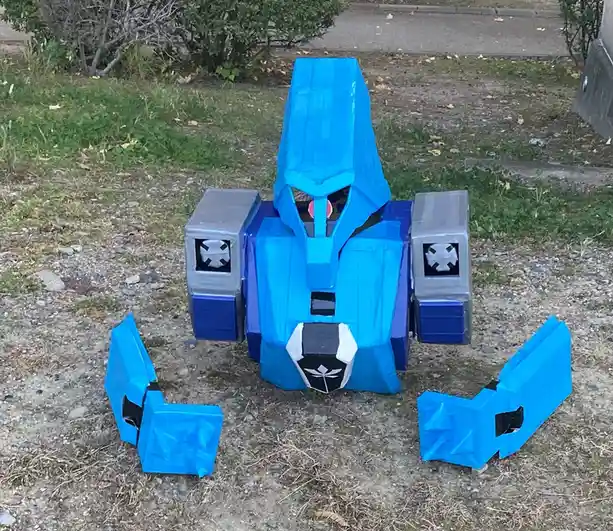 A homemade robot costume made of blue and gray duct tape sits on a gravel and grass patch outdoors, with its two arm pieces…
