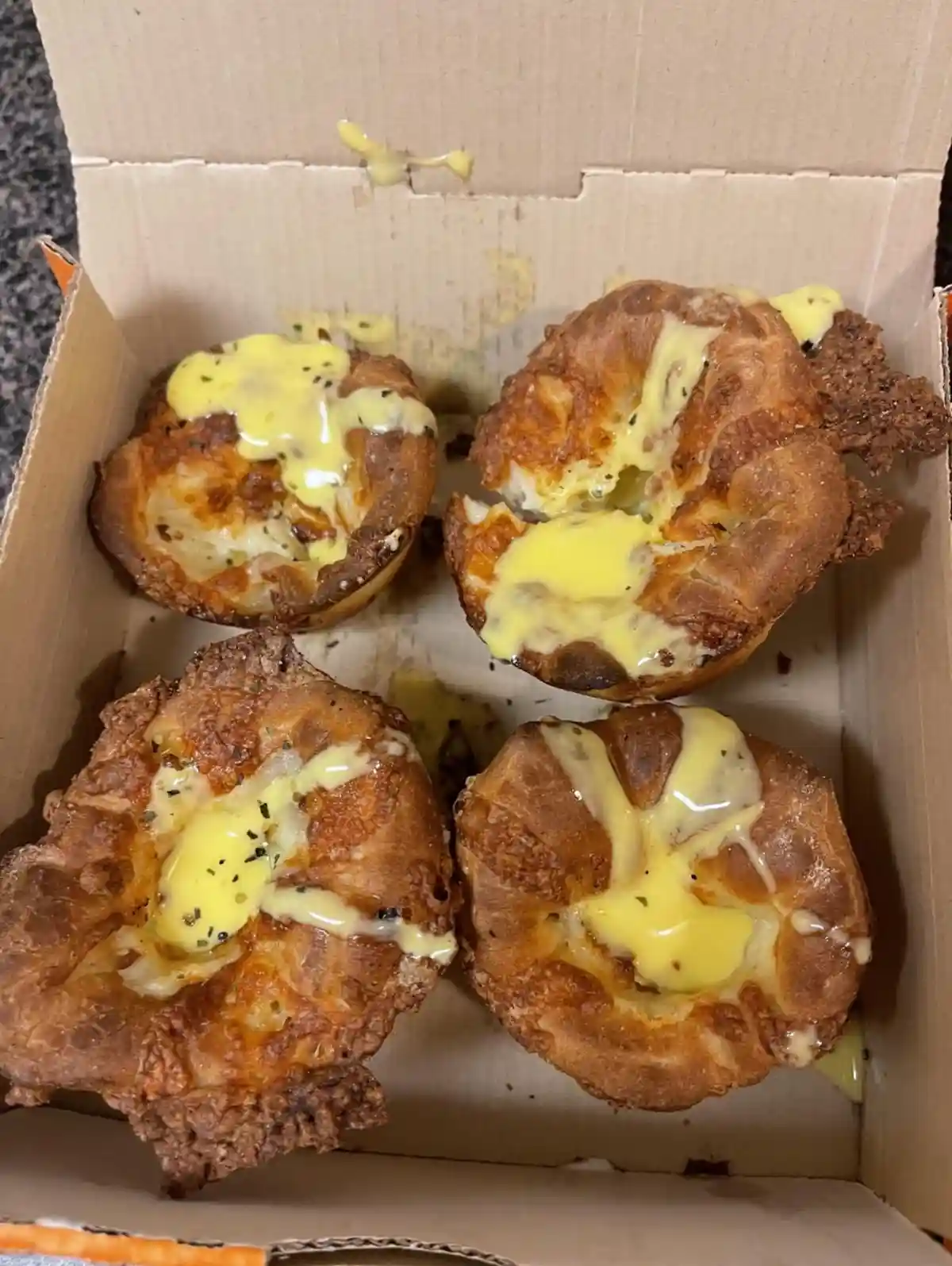 A cardboard box contains four golden-brown Yorkshire puddings, each topped with a generous amount of yellow sauce and…