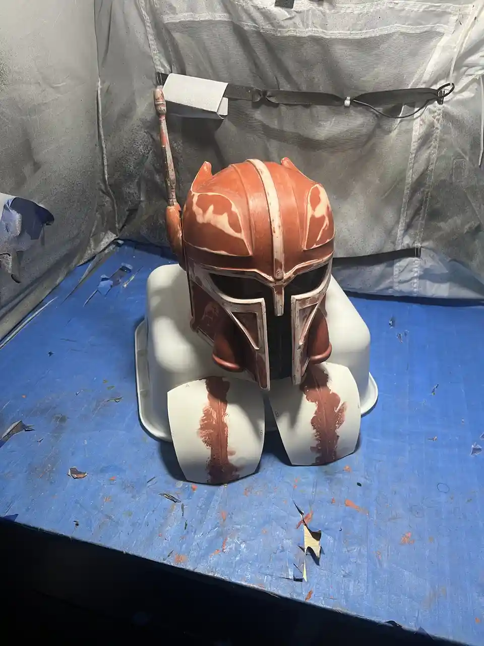 A Mandalorian-style helmet, likely Boba Fett's, sits on a white stand inside a spray-paint booth with blue plastic sheeting…