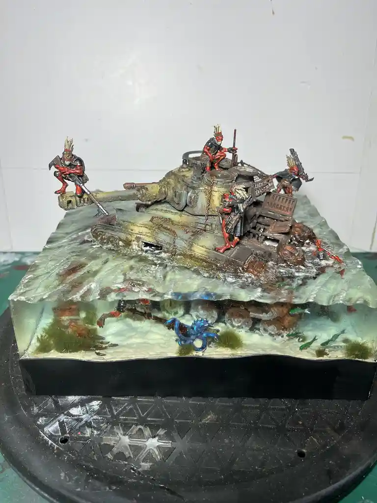 A detailed miniature diorama shows a heavily weathered tank partially submerged in clear resin, with red-skinned, horned…
