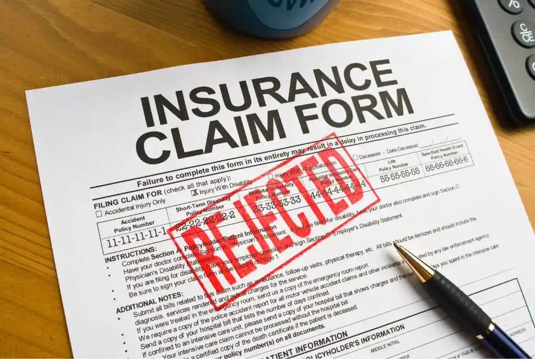 A close-up photo of an insurance claim form on a wooden desk, with a large red 'REJECTED' stamp across the middle. A pen…