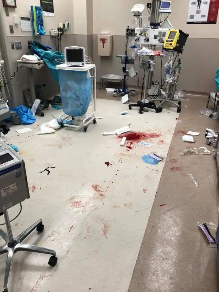 A messy hospital room with medical equipment, scattered papers, and noticeable bloodstains on the floor. The overlaid text…
