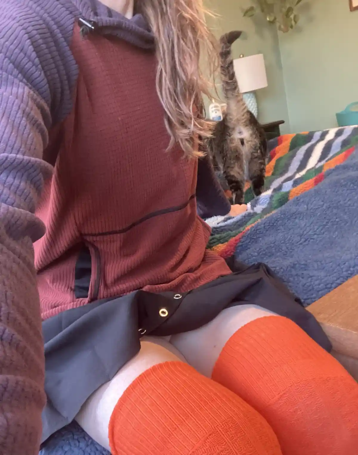 A person wearing a purple hoodie, red vest, and orange tights sits on a bed with a colorful blanket. A tabby cat walks away…