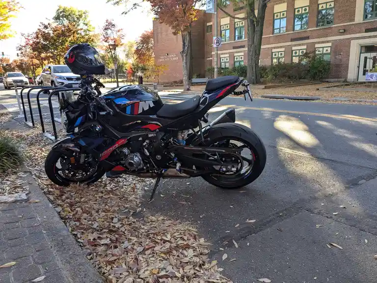 A black BMW S1000RR motorcycle with red and blue accents is parked on a street covered in autumn leaves, with a black helmet…