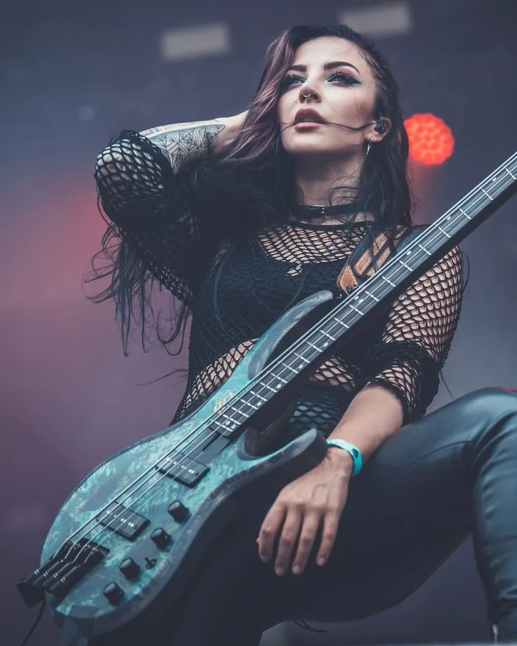 A woman with dark hair and tattoos, wearing a black fishnet top and black pants, sits confidently holding a teal bass…