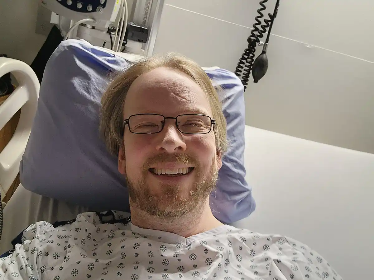 A man with glasses and a beard, wearing a hospital gown with a snowflake pattern, is lying in a hospital bed with medical…
