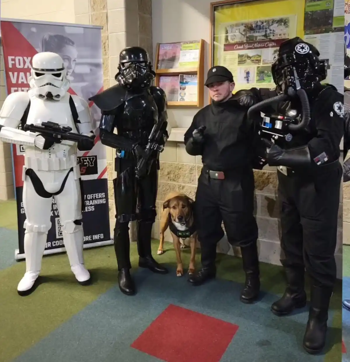 Four people dressed as Star Wars stormtroopers and an Imperial officer pose indoors with a brown dog wearing a harness. One…