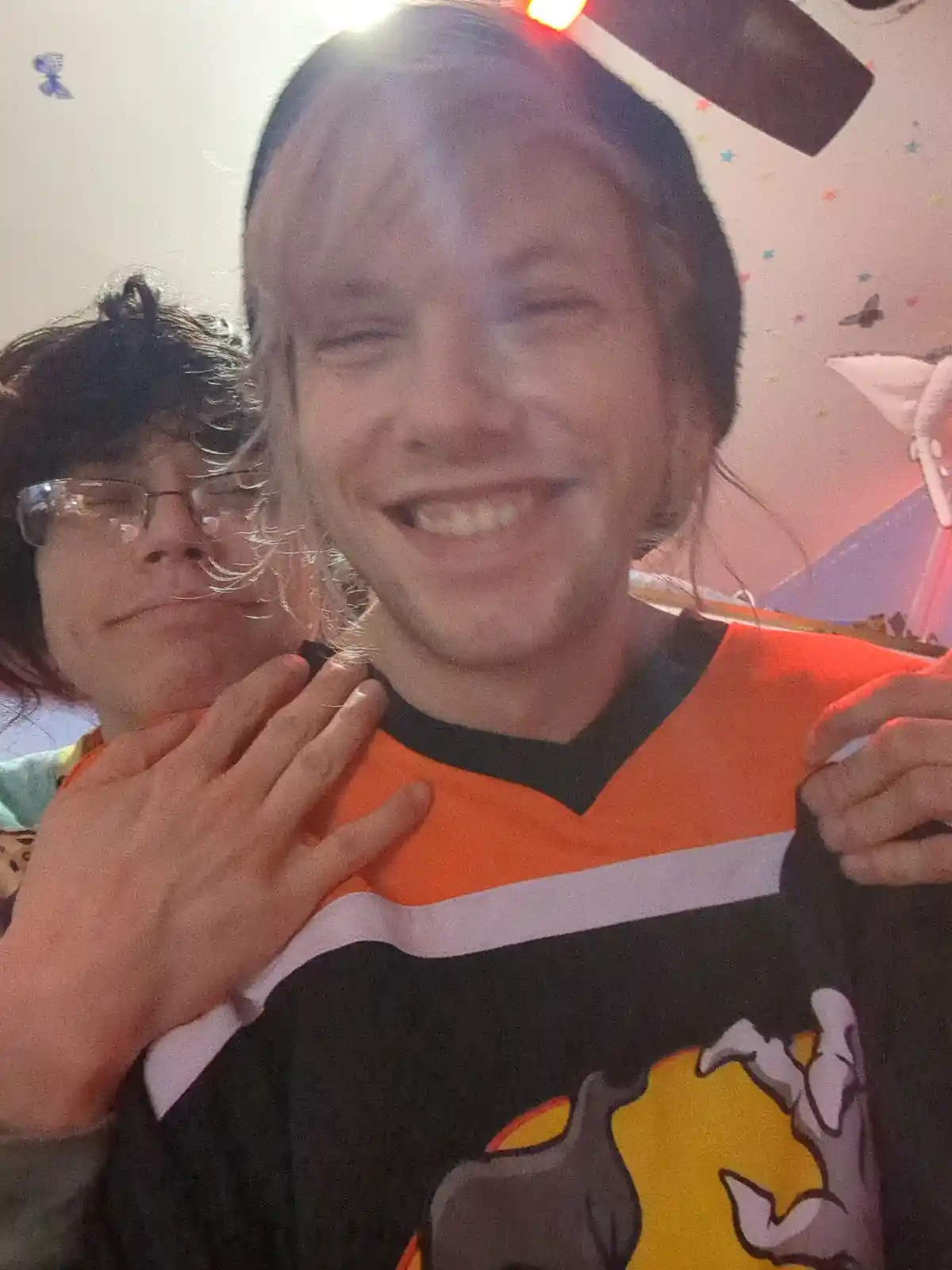 A person with long blonde hair and a wide, joyful smile wears an orange, black, and white hockey jersey with a cartoon…