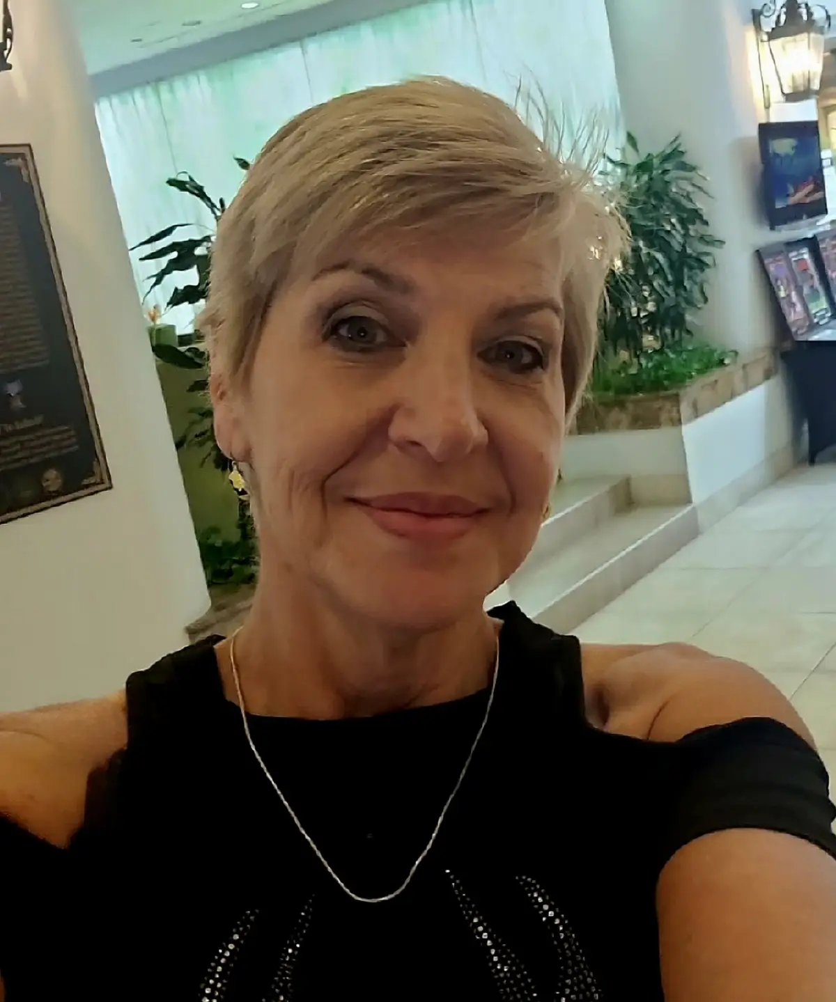 A selfie of a smiling woman with short blonde hair, wearing a black top and a silver necklace, taken indoors in a well-lit…