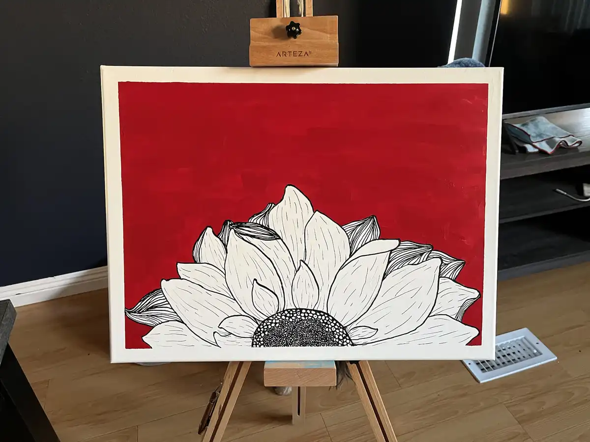 A painting of a white sunflower with black outlines and a black dotted center is displayed on a wooden easel against a red…