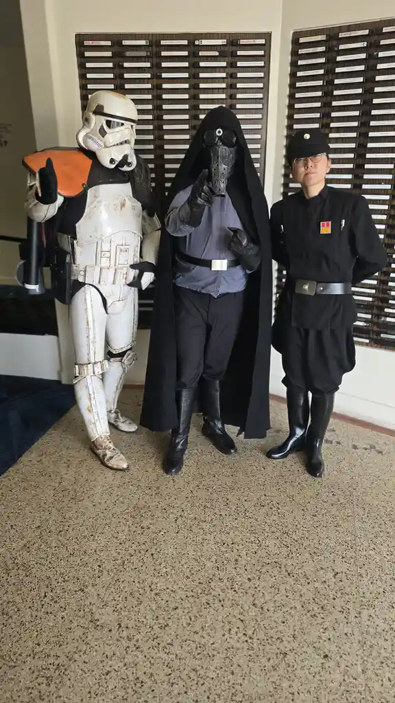 Three people pose indoors in front of a wall of VHS tapes, dressed as Star Wars characters: a dirty white stormtrooper, a…