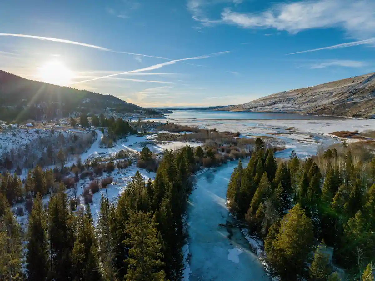 An aerial photo shows a frozen river winding through a snowy forest with mountains in the background under a bright sun. The…