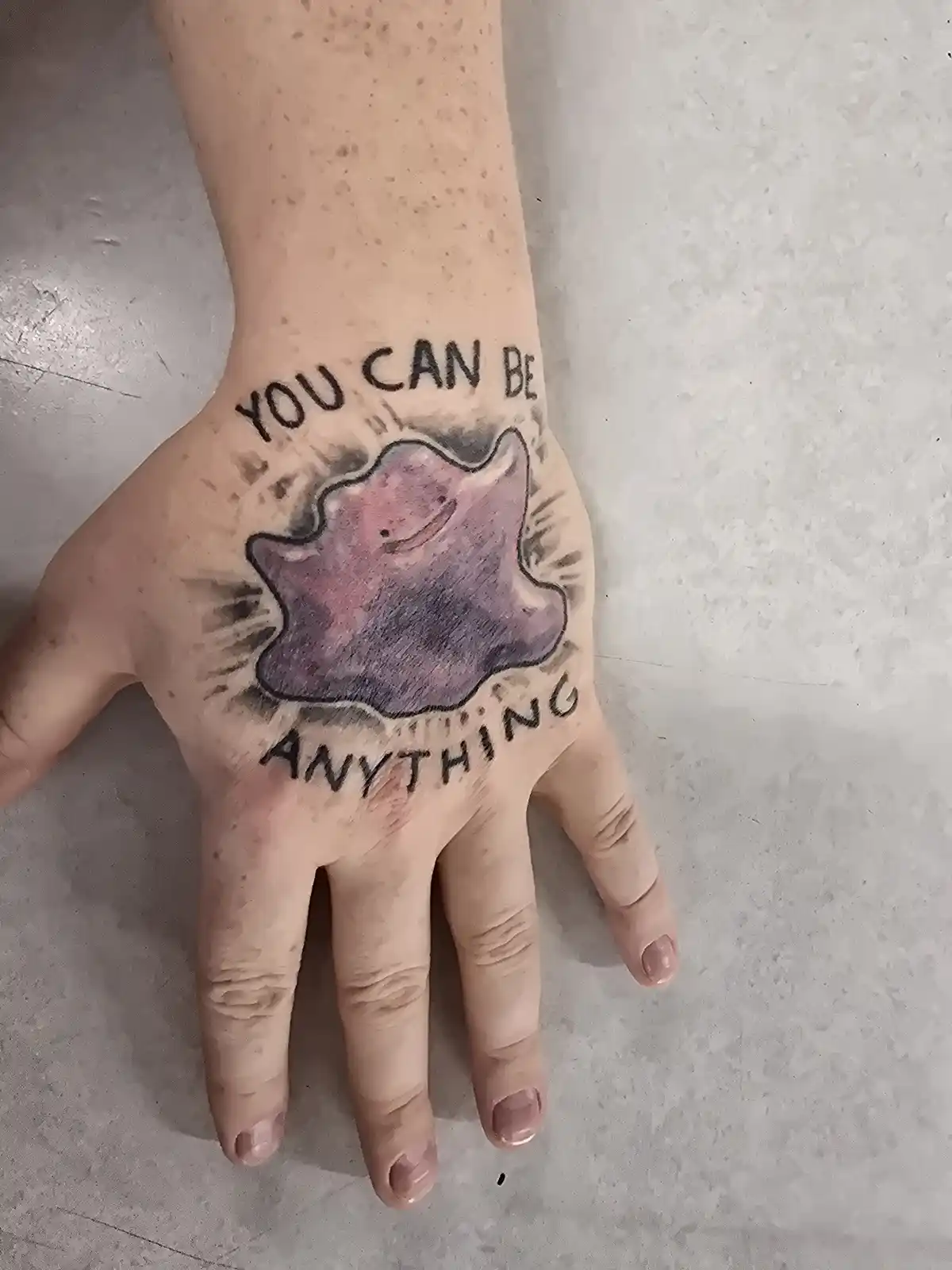 A close-up photo shows a person's open palm with a tattoo of the Pokémon Jigglypuff, colored in purple and pink with a…